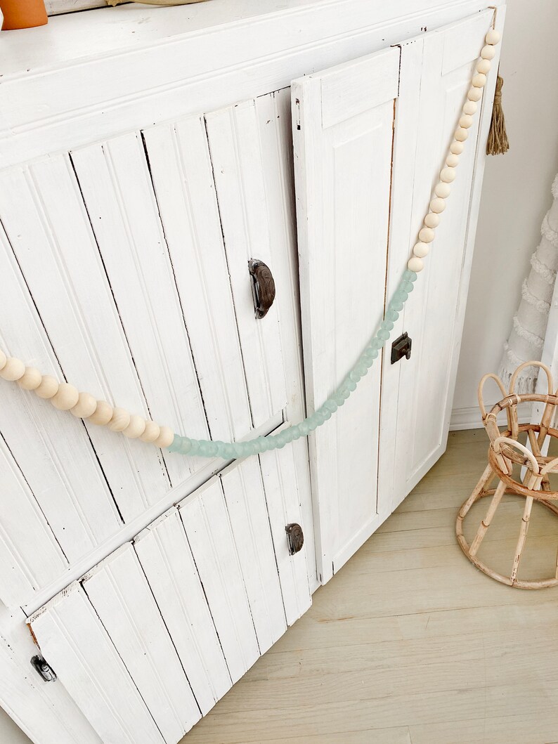May include: White wooden cabinet with two doors and beaded garland draped across the front. The garland is made of light brown and light blue beads.