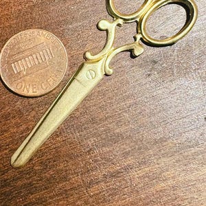 May include: A small, gold-toned metal scissor charm featuring ornate, curved handle loops and a single, non-functional blade design. The item is positioned next to a copper-colored United States one-cent coin for scale, highlighting its miniature size.