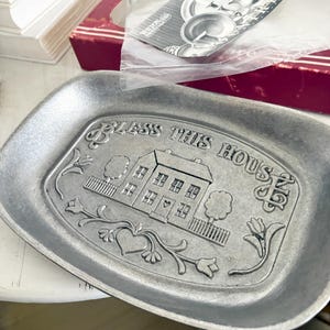 May include: A silver-colored metal tray with the embossed phrase "BLESS THIS HOUSE" and a detailed image of a house, trees, and floral accents. Additional silverware is visible in the background.