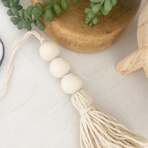 May include: A white wooden bead garland with a cream-colored tassel. The garland has three beads and is hanging from a white string.