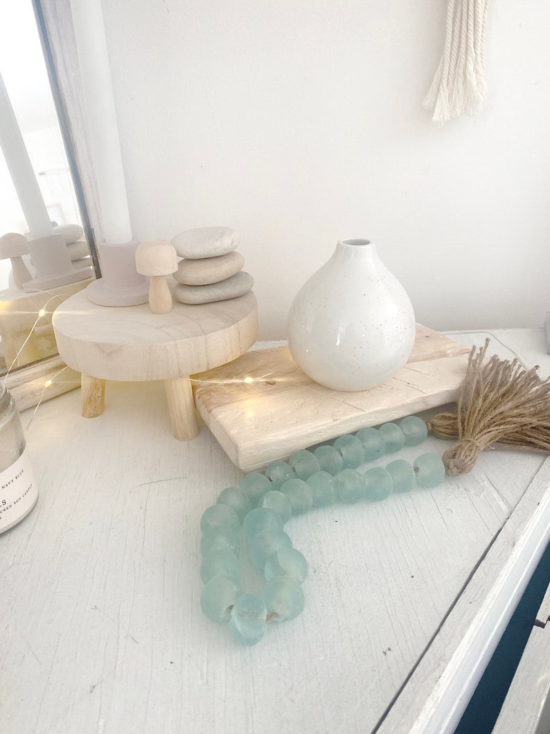 Op de afbeelding: Een witte keramische vaas met een gespikkelde afwerking staat op een houten snijplank met een lichtblauwe glazen kralenkrans en een bruine kwast.