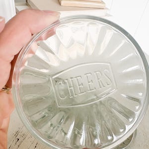 May include: Clear glass coaster with a fluted design and the word "CHEERS" embossed in a rectangular frame. The coaster is held in a hand, with a blurred background.