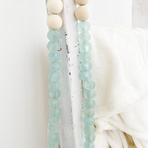 May include: A light blue glass bead garland with white wooden beads. The garland is draped over a white wooden ladder.