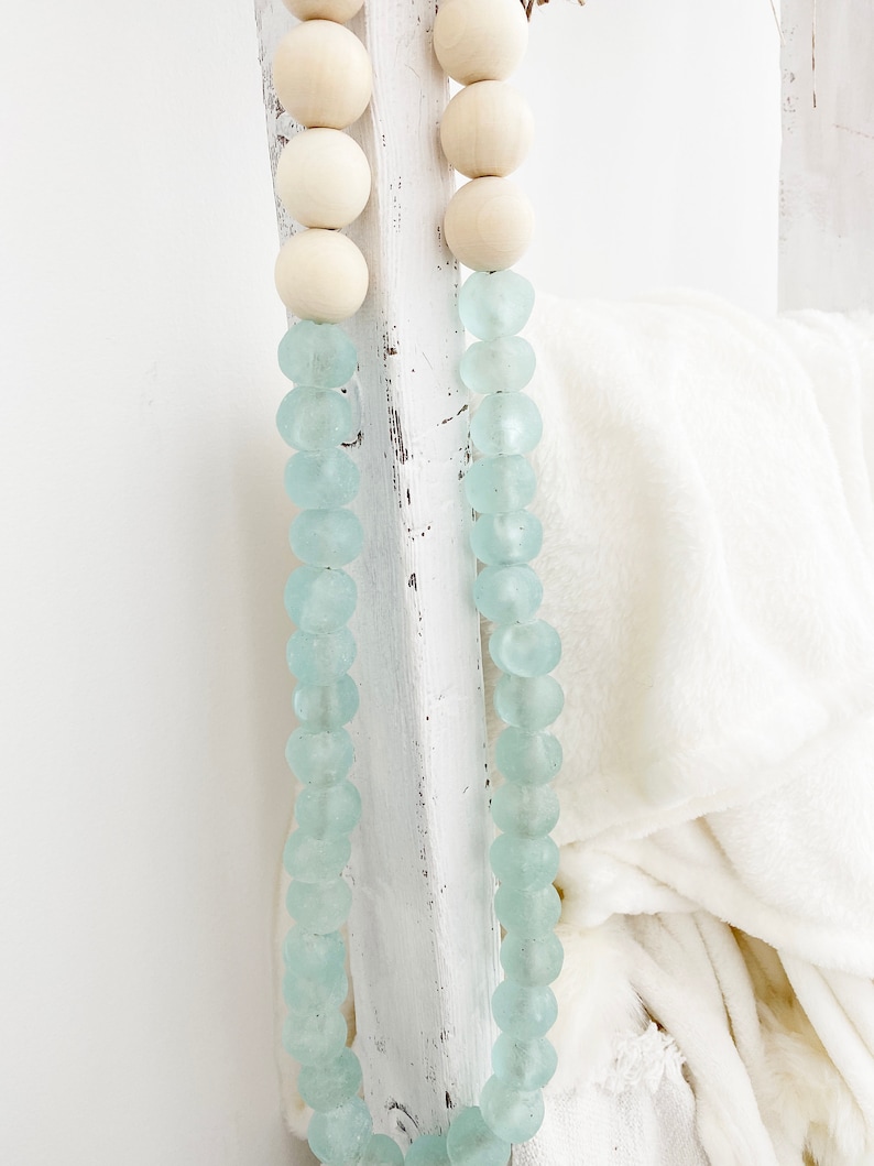 May include: A light blue glass bead garland with white wooden beads. The garland is draped over a white wooden ladder.