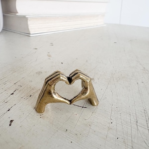 May include: A small, gold-colored metal sculpture of two hands forming a heart shape. The sculpture is on a distressed, off-white surface, with a stack of books in the background. The hands are detailed, with visible fingers and a textured finish.