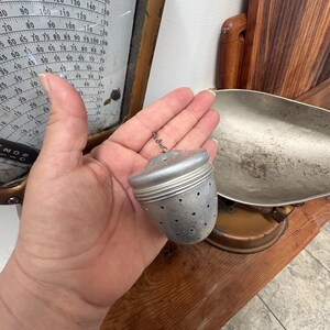 May include: A silver-colored metal tea infuser with a perforated, acorn-shaped body and a small attached chain. The infuser features a threaded lid design and numerous small circular holes distributed across the surface for steeping loose tea leaves.