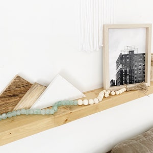 May include: A wooden shelf with a white and light brown geometric design, a black and white photo of a building, and a light blue and white beaded garland.