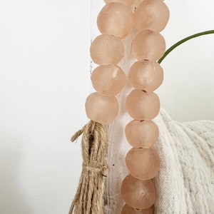 May include: A strand of light pink glass beads with a natural fibre tassel. The beads are round and have a slightly frosted finish.