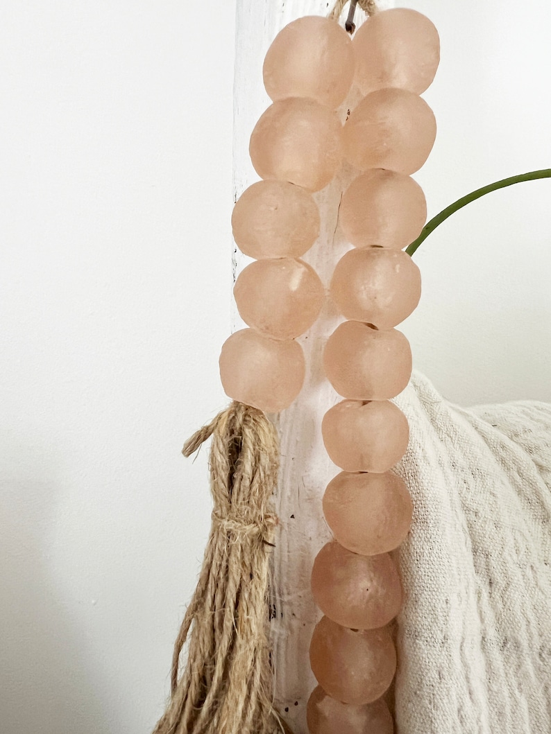 May include: A strand of light pink glass beads with a natural fibre tassel. The beads are round and have a slightly frosted finish.