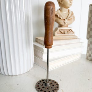 May include: A vintage potato masher with a wooden handle and a circular, perforated metal head. The handle is a dark brown color, and the metal head shows signs of age. The masher is standing upright on a white surface.