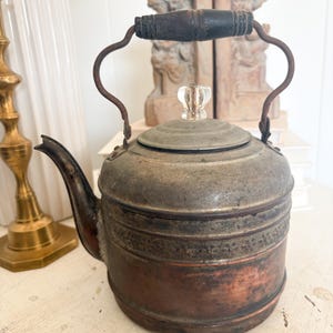 May include: A vintage metal kettle featuring a weathered silver-toned upper body and a warm copper-toned lower section. It includes a dark wooden handle, a curved spout, and a lid topped with a clear glass knob, showcasing a decorative floral band.