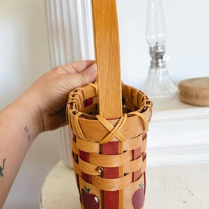 May include: A woven basket with a wooden handle. The basket is made of light brown and red woven strips, with a repeating apple design. The handle is a single piece of light brown wood.