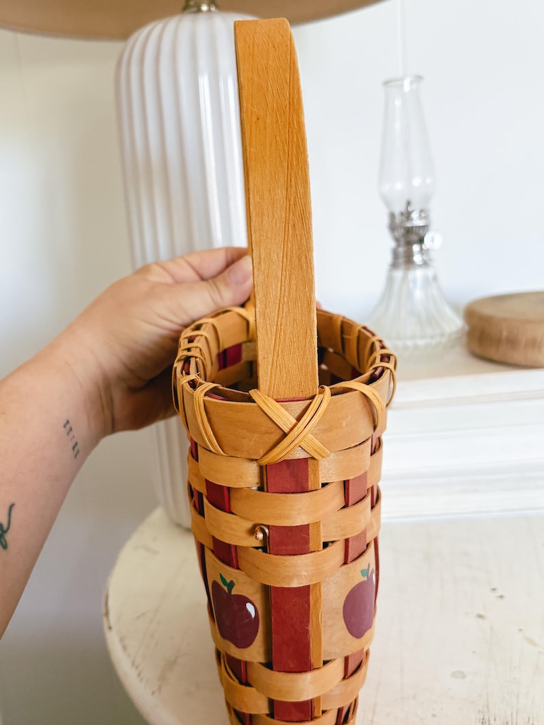 May include: A woven basket with a wooden handle. The basket is made of light brown and red woven strips, with a repeating apple design. The handle is a single piece of light brown wood.