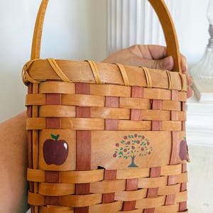 May include: A woven basket with a wooden handle. The basket is made of light brown and red woven strips. It features painted images of a red apple and a tree with red apples.