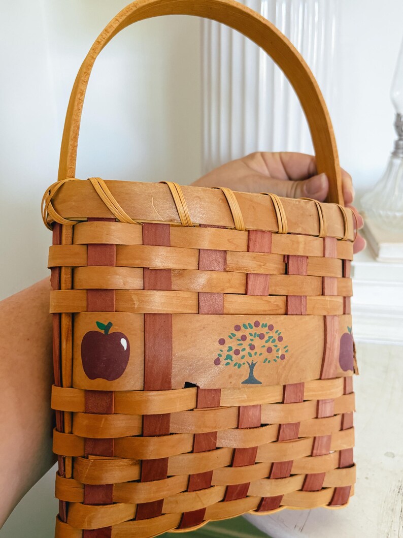 May include: A woven basket with a wooden handle. The basket is made of light brown and red woven strips. It features painted images of a red apple and a tree with red apples.