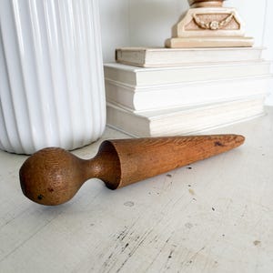 May include: A wooden rolling pin with a rounded handle and a tapered rolling surface. The rolling pin is made of a light brown wood and sits on a white surface. A white vase and a stack of books are in the background.