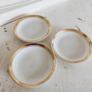 May include: Three small, round, white ceramic dishes with a gold rim. The dishes are arranged on a distressed white surface. A stack of white books is in the background.