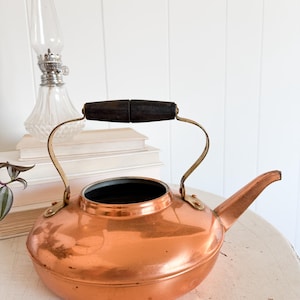 May include: A copper kettle with a dark wooden handle and brass accents. The kettle has a spout and a rounded body. A clear glass oil lamp and a stack of books are in the background. The kettle is on a white surface.
