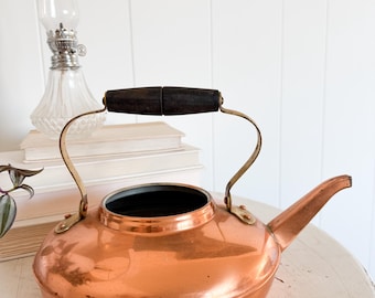 May include: A copper kettle with a dark wooden handle and brass accents. The kettle has a spout and a rounded body. A clear glass oil lamp and a stack of books are in the background. The kettle is on a white surface.
