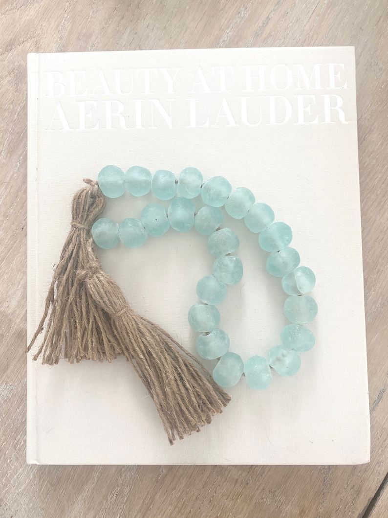 Op de afbeelding: Een lichtblauwe glazen kralenketting met een bruine jute kwast. De ketting ligt op een wit boek met de tekst "BEAUTY AT HOME AERIN LAUDER".