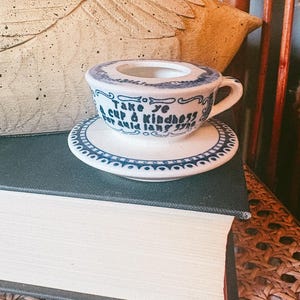 May include: A small white ceramic cup and saucer set with blue decorative trim. The cup features the black text "Take ye a cup o kindness for auld lang syne" written on the front. The saucer has a matching blue patterned border design.