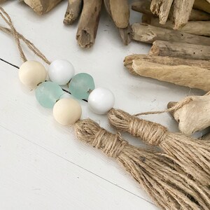 May include: A handmade macrame wall hanging with two tassels. The wall hanging features a string of beads with white, light blue, and beige beads.