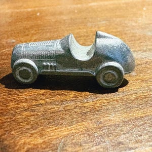 May include: A small, silver-colored metal toy race car featuring an open cockpit, a textured hood with horizontal lines, and four wheels. The vehicle has a streamlined, vintage-inspired body shape with a rounded rear section and a low profile.