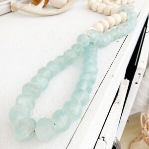 May include: A light blue glass bead garland with a white background. The garland is made of round, frosted glass beads.
