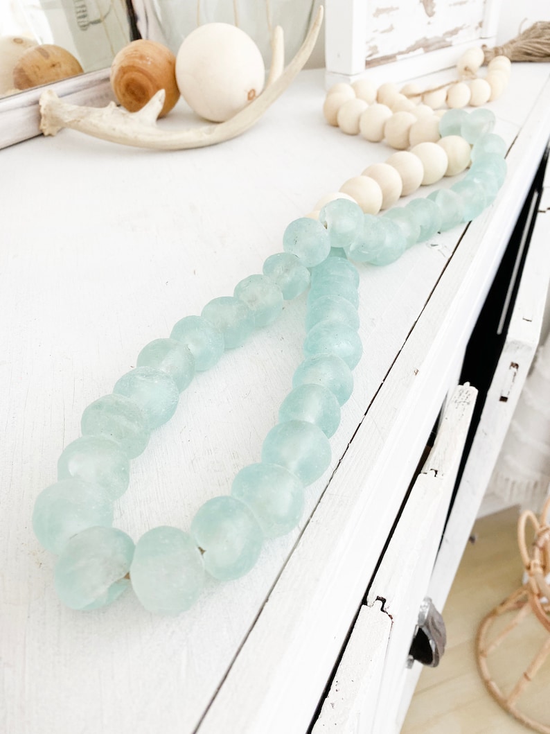 May include: A light blue glass bead garland with a white background. The garland is made of round, frosted glass beads.