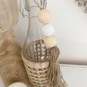 May include: A clear glass bottle with a woven straw cover and a brown tassel. The bottle has a white and beige beaded garland attached to the top.
