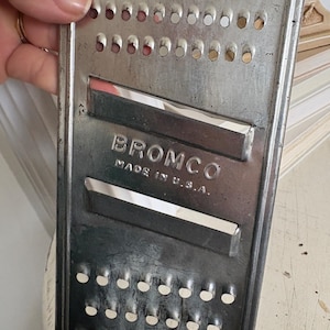 May include: A vintage metal grater with the brand name "BROMCO" and the text "MADE IN U.S.A." stamped on the surface. The grater has multiple rows of small, round holes and two rectangular openings. The metal has a worn, aged appearance.
