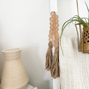 May include: A white wooden ladder with a pink glass bead garland with brown tassels hanging from it. A white blanket is draped over the ladder. A small potted plant is on the top shelf of the ladder.