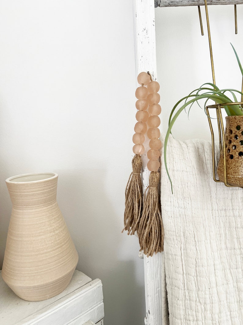 May include: A white wooden ladder with a pink glass bead garland with brown tassels hanging from it. A white blanket is draped over the ladder. A small potted plant is on the top shelf of the ladder.