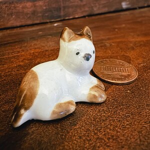 May include: A small ceramic figurine of a cat in a lying position with white and brown patches. The cat has pointed ears, black eyes, and a dark nose. It is positioned next to a copper-colored United States one-cent coin for scale reference.