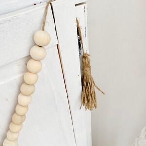 May include: A white wooden cabinet door with a wooden bead garland and a brown jute tassel hanging from the top.