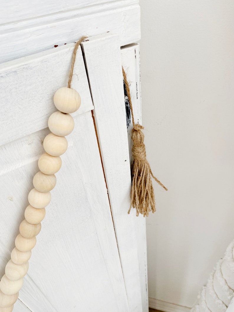 May include: A white wooden cabinet door with a wooden bead garland and a brown jute tassel hanging from the top.