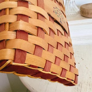 May include: Woven basket with a rectangular shape, featuring a natural wood color and burgundy accents. The basket has an apple and tree design. The basket is sitting on a white surface.