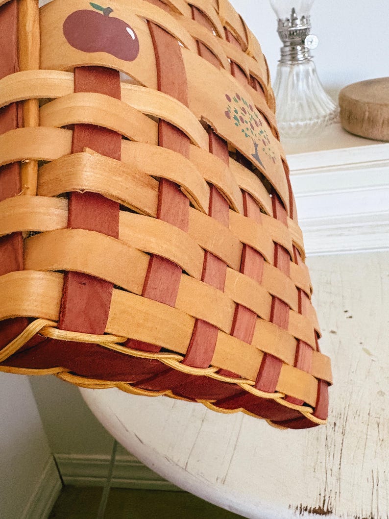 May include: Woven basket with a rectangular shape, featuring a natural wood color and burgundy accents. The basket has an apple and tree design. The basket is sitting on a white surface.