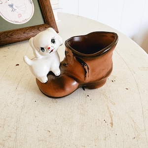 May include: A vintage ceramic planter shaped like a brown boot with a white puppy figurine perched on top. The puppy has black eyes and a red nose. The planter is on a white surface.