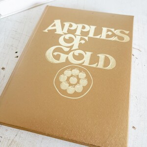 May include: A hardcover book with a gold cover and gold lettering that reads "Apples of Gold." The book has a circular design of apples in the center. The book is lying on a white surface.