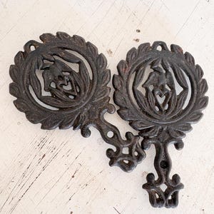 May include: Two antique cast iron trivets. Each trivet features a circular design with a central figure and a surrounding leaf pattern. The trivets are connected by an ornate handle. The color is a dark, aged brown.