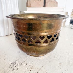 May include: A vintage brass planter with a heart-shaped cut-out design around the base. The planter has a wide, rounded bowl shape and a slightly flared rim. The brass has a warm, aged patina.