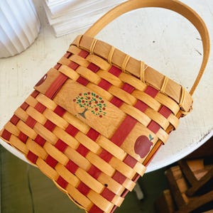 May include: A woven basket with a wooden handle. The basket features a red and natural wood weave pattern. A wooden panel on the front is decorated with a tree and apple illustrations. The basket is designed for carrying items.