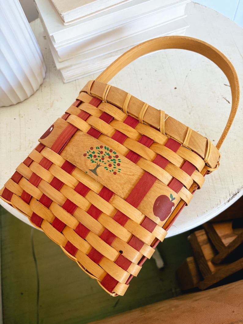 May include: A woven basket with a wooden handle. The basket features a red and natural wood weave pattern. A wooden panel on the front is decorated with a tree and apple illustrations. The basket is designed for carrying items.