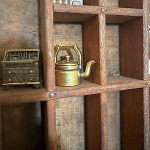 May include: A wooden printer's tray with multiple compartments containing miniature items, including a small brass-colored metal teapot, a dark metal desk with drawers, a set of miniature books, and a tall, thin vertical metal object.