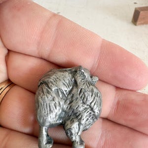 May include: A small, silver-toned metal figurine depicting the rear view of a long-haired dog. The sculpture features detailed, textured fur patterns across the back and hindquarters, with two visible legs standing on a flat surface.