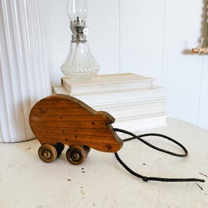 May include: A vintage wooden pull toy shaped like a bear, with four wooden wheels and a black rope. The bear is made of stained wood. A clear glass oil lamp and a stack of books are in the background.