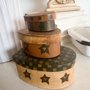 May include: A stack of four oval-shaped decorative boxes. The boxes are painted in shades of brown, tan, and green. The largest box has a tan base with black star accents. The next box has a brown base with a green and black checkered top.