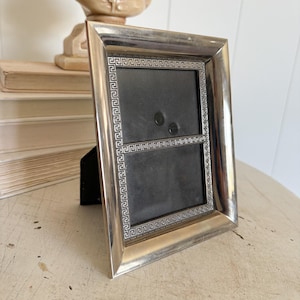 May include: A rectangular silver-toned metal picture frame featuring a polished outer border and an inner border decorated with a repeating Greek key pattern. The frame has a dark gray backing and a black easel stand attached to the rear side.