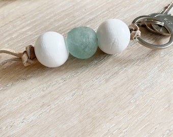 May include: A white and light blue beaded keychain with a silver key ring. The beads are round and smooth, and the keychain is made of a brown leather cord.